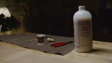 a set of leatherworking tools, including a spool of thread, awl, pliers, and a bottle of finishing solution, arranged on a brown suede mat atop a wooden workbench.
