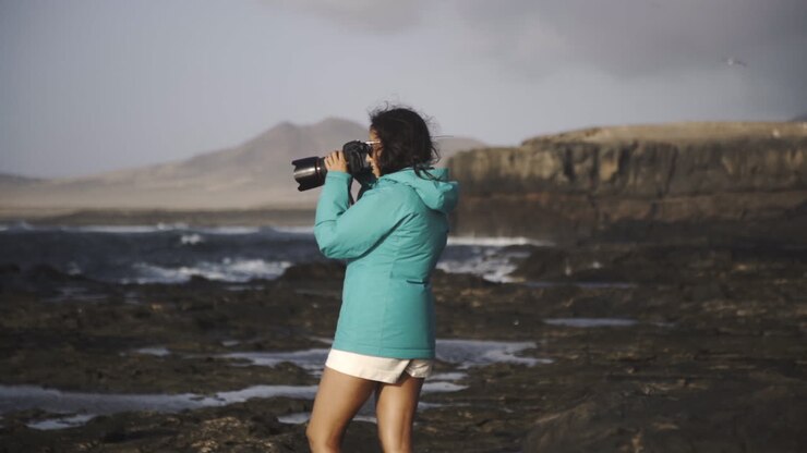 freelancer creative photographer standing on ocean cliff during a windy day wearing trendy jacket taking photos of natural seascape in Fuerteventura Spain canary island