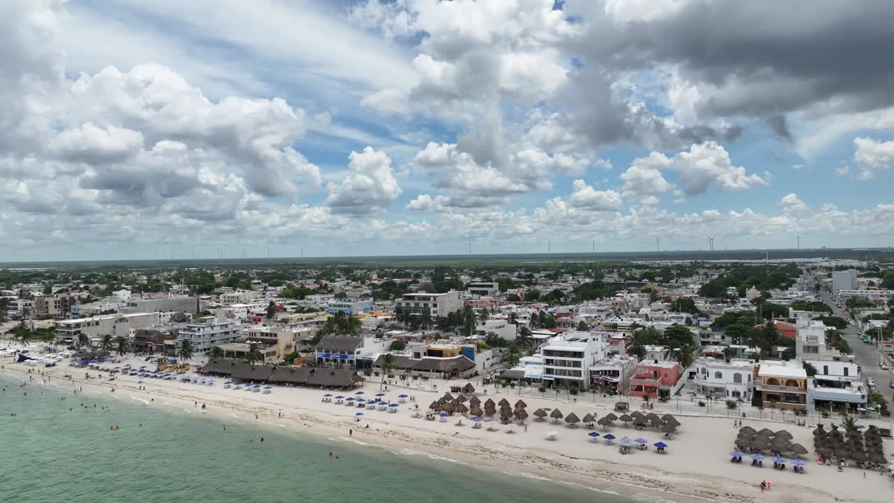 A lateral drone flight over Puerto Progreso, showing the beachfront city with crowded sandy shores, turquoise water, and dramatic towering clouds on a bright sunny day