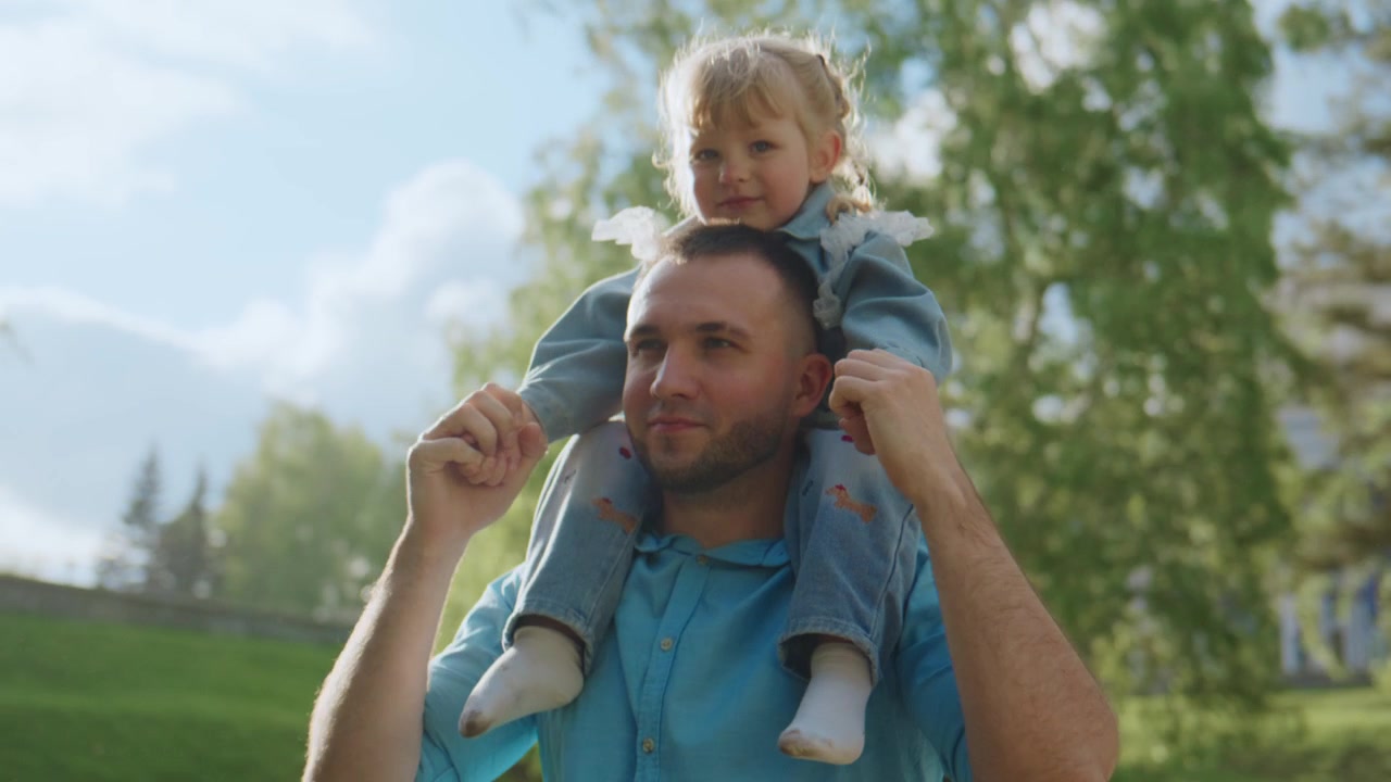 Pai carrega a filha pequena nos ombros ao ar livre. Ela segura a cabeça dele com as duas mãos, sorrindo. Ele olha para frente, aproveitando o momento deles. Sol forte ilumina árvores verdes e grama