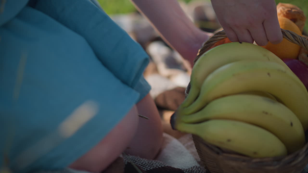 una toma de primer plano de la mano de una mujer, con un vestido azul fluido, mientras se inclina para colocar una canasta llena de plátanos, manzanas y otras frutas en una manta de picnic a cuadros en un campo cubierto de hierba