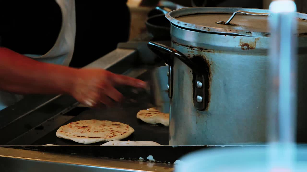 A close up view of a woman flipping several mexican tortilla on a grill. Huge pot in the foreground with beans cooking inside. Customers waiting for the food to be placed in a box.