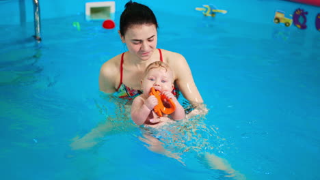positive woman is moving her baby in the swimming pool. little infant boy is chewing a toy. teaching a child to swim from birth.