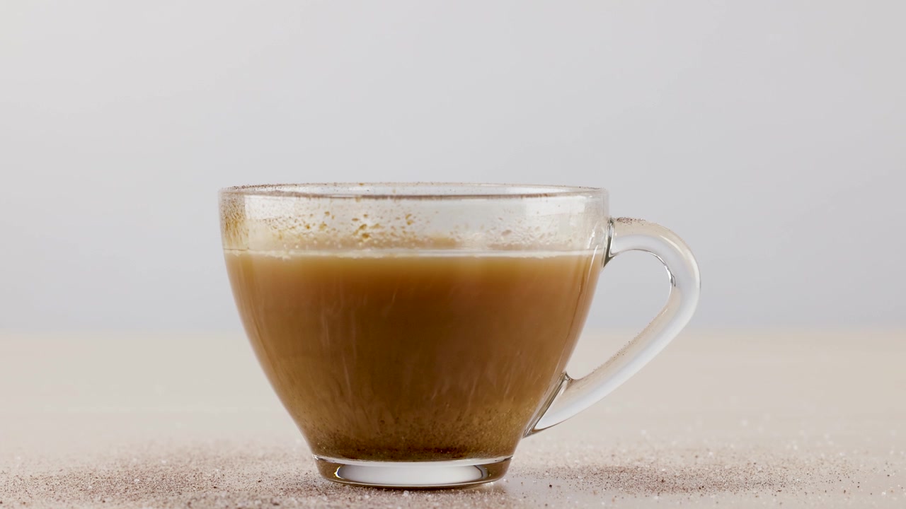 Zucchero granulato cade in una tazza di caffè in vetro su uno sfondo bianco pulito.
