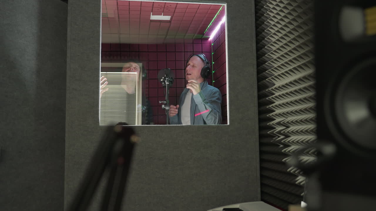 Two men in a red soundproof room sing passionately. One wears a black cap and shirt, and the other is dressed in a blue shirt and white inner shirt. Both are engaged in a focused, musical performance