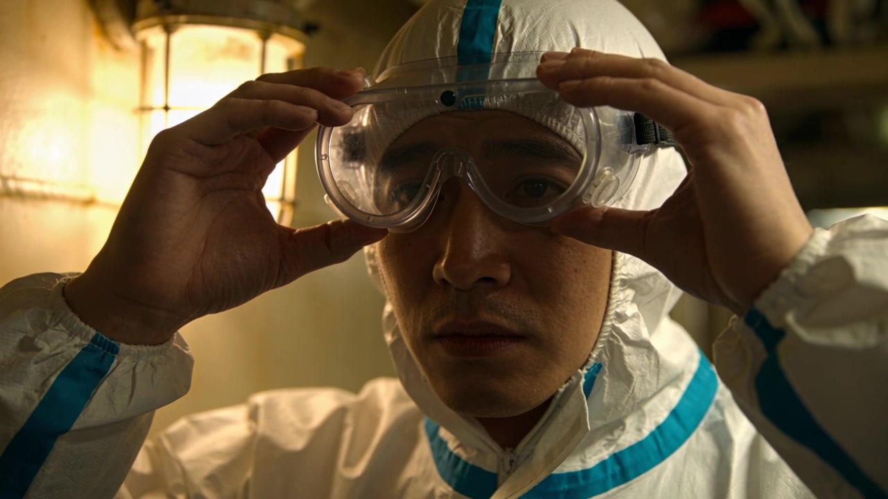 Uomo in piedi in tuta bianca con cuciture nastrate in blu e occhiali protettivi trasparenti in un'area di laboratorio, che si prepara per un compito.
