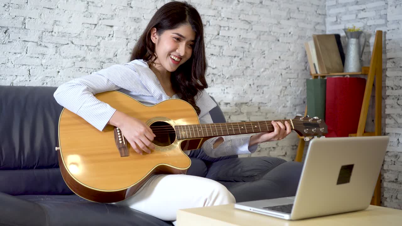 Joven mujer asiática practicando y aprendiendo a tocar la guitarra en la pantalla de un portátil. Guitarrista viendo un tutorial en línea.