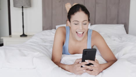 Happy-biracial-woman-lying-on-bed-using-smartphone,-slow-motion