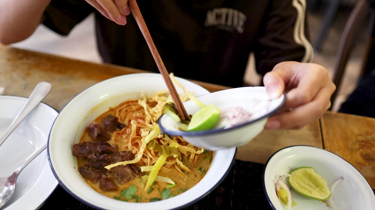 A person squeezes fresh lime over a bowl of beef curry noodles in a casual Thai restaurant, with natural lighting and close-up camera angles