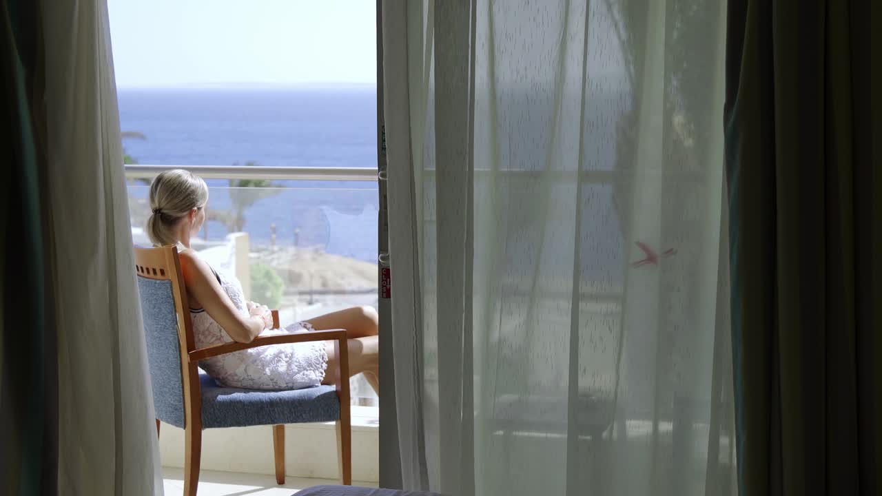 una mujer joven se sienta en un balcón de un hotel y disfruta de una hermosa vista del mar y las palmeras.