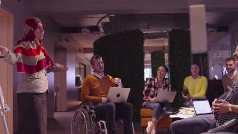muslim woman in a modern open space coworking office giving a presentation to team. middle eastern arab businesswoman in creative startup office. disabled businessman in a wheelchair on team meeting