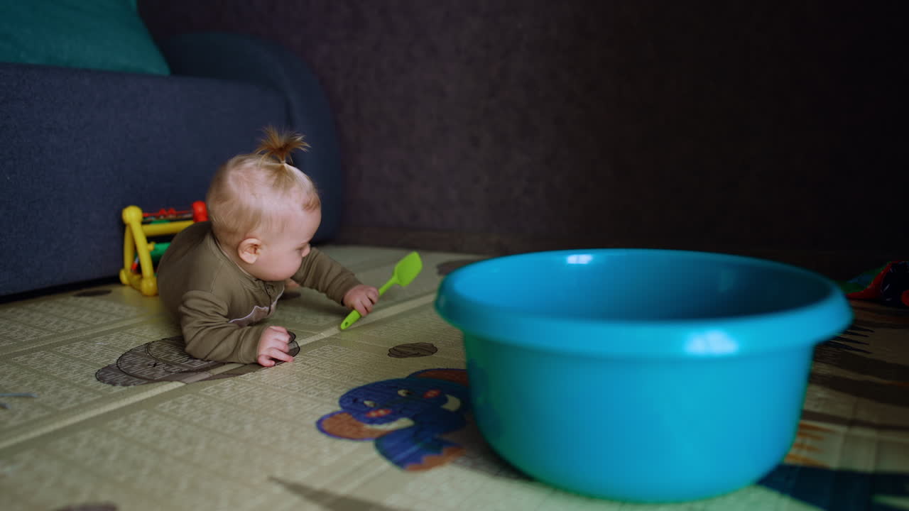 Small kid with a shovel in hands lies on the floor. A blue washtub is near the infant.