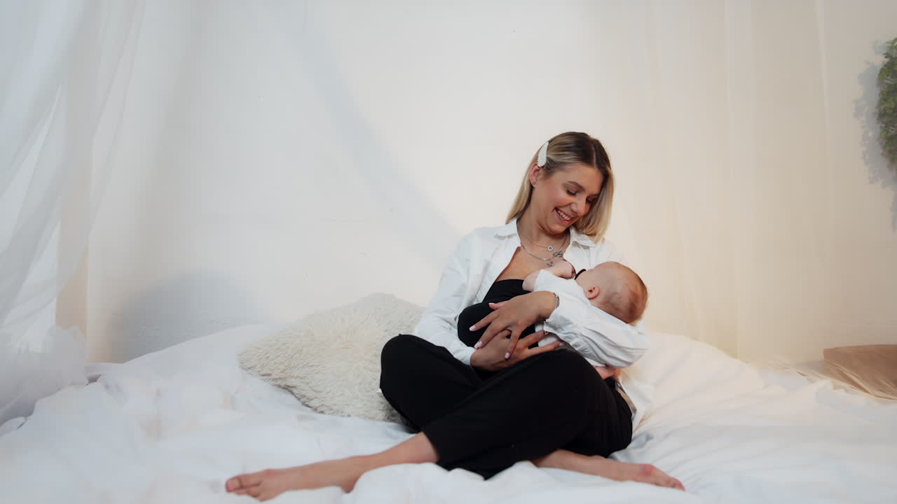 Laughing blonde woman sits on the bed holding a baby in her arms. Mother and child portrait.