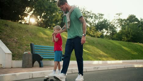 A-father-in-a-green-T-shirt-and-blue-jeans-helps-his-son-in-a-red-T-shirt-and-blue-shorts-ride-a-skateboard-along-the-road-in-the-park.-Happy-father-and-son-spending-time-active-leisure