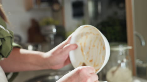 chef in green outfit pours brown mashed ingredient from white bowl into stainless pot during meal preparation in rustic kitchen with blurred background