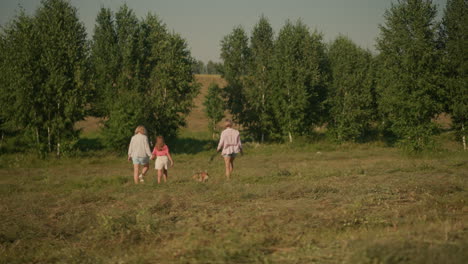 back view of dog owner walking dog on leash and mother holding her daughter's hand as they stroll together through farmland with lush greenery and distant trees under clear blue sky