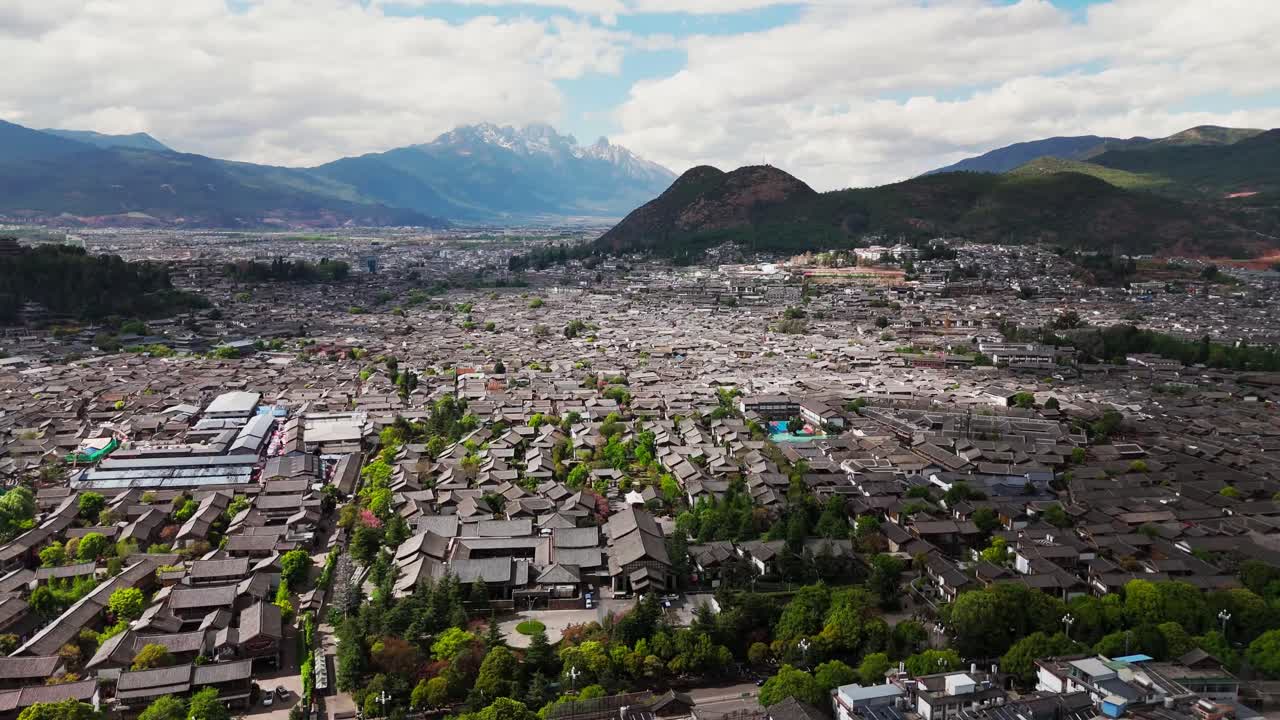 Wide aerial of Lijiang Old Town framed by open sky and forested mountain slopes, panoramic establishing dolly