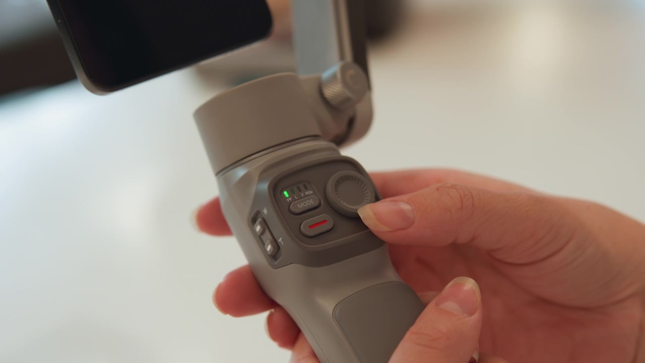 Tight front-right close-up of hands operating handheld gimbal control panel, thumb pressing directional pad while index finger hovers near mode buttons, showing precise adjustment, steady handling