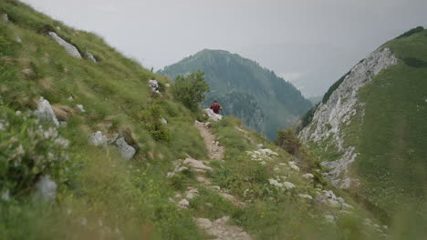 A-hiker-in-red-jacket-walking-with-hiking-poles-on-a-narrow-path-on-a-mountain-towards-the-camera-in-the-distance-are-mountains-in-low-cloud-cover