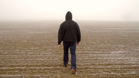 Caucasian-male-in-hoodie-walking-alone-in-arid-field-during-mysterious-thick-fog,-slow-motion-follow-shot