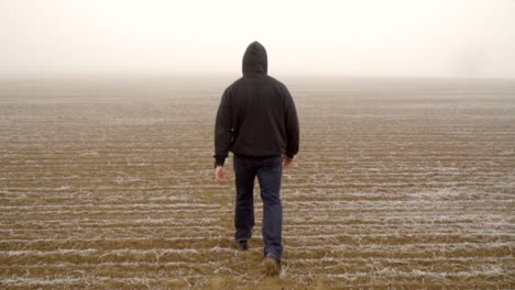 caucasian male in hoodie walking alone in arid field during mysterious thick fog, slow motion follow shot