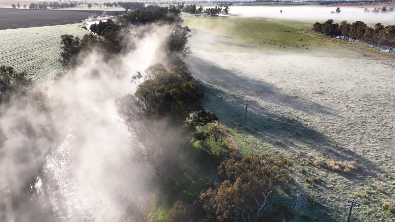 Drone footage glides above a frosty meadow and tree line, capturing early morning fog illuminated by low, golden sunlight in a rural mountain landscape