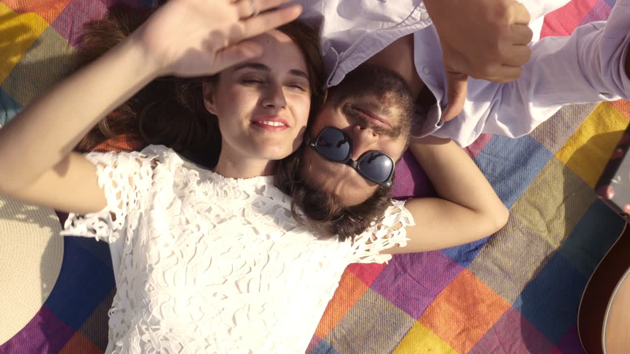 행복하고 사랑스러운 젊은 부부는 공원에서 다채로운 담요에 누워서 셀카를 찍습니다. 기타, 선글라스와 함께 만적인 스마트폰. 아름다운 매력적인 소녀.  뷰. 회전하는 카메라. 느린 동작.