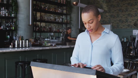 Female-Owner-Of-Restaurant-Bar-Standing-By-Counter-Checking-Reservations-Before-Service