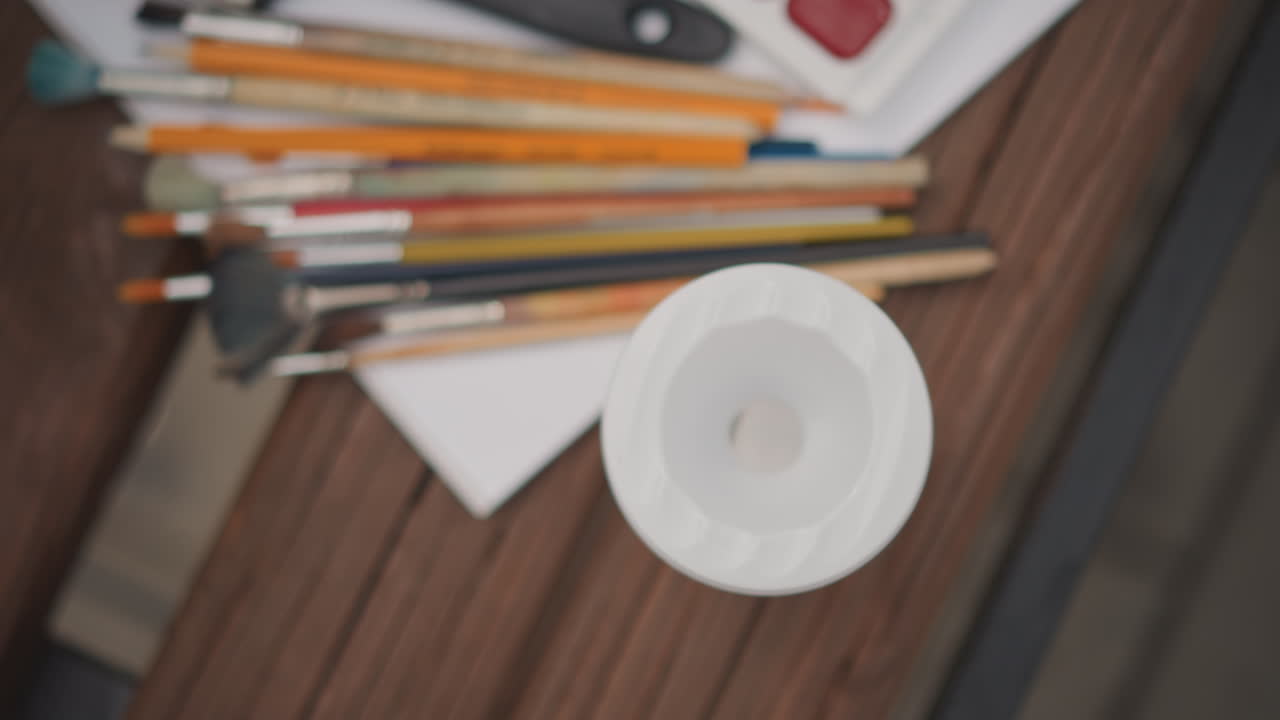 Instructor demonstrates painting rinsing process on table. Topdown shot of art tools and organized supplies scene. Still image showing art instructor teaching rinsing techniques over cluttered table