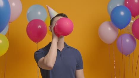 studio portrait of man wearing party hat celebrating birthday blowing up balloon 1