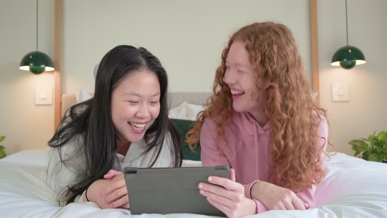 Due amiche sdraiate sul letto, che reagiscono a una clip sul tablet, toccano lo schermo e ridono in camera da letto