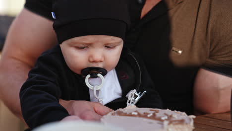 beautiful baby with pacifier wearing black cap. close up. little infant sitting in dad's hands touching a cake. close up.