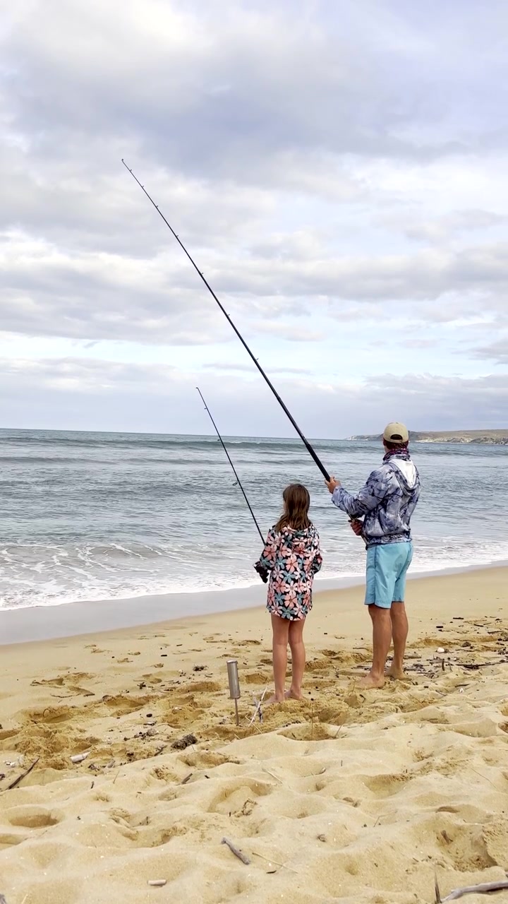 Papà e figlia con le canne da pesca in mano, che pescano da una spiaggia sabbiosa nell'oceano. Verticale.