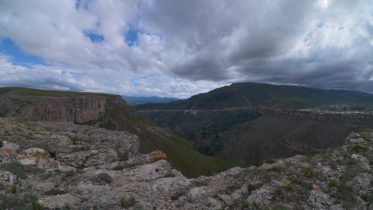 Un time-lapse cattura nuvole drammatiche che si muovono sopra una vasta valle montuosa verde con scogliere rocciose e vette distanti sotto un cielo mutevole.