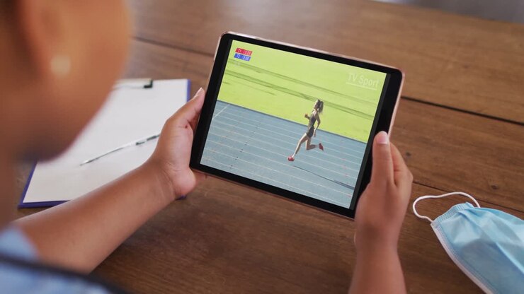 Composite of woman sitting at table with face mask, watching athletics running event on tablet