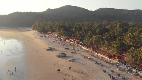 low tide palolem beach at sunset with tourists enjoying the paradisiac ocean shore, in goa, india- aerial low angle slow orbit shot