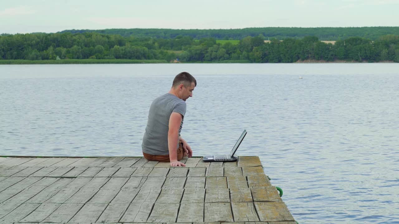 Handsome man sits on a wooden pier with laptop. Work on vacation
