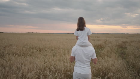 Vista-Desde-Atrás:-Papá-Lleva-Sobre-Sus-Hombros-A-Su-Querida-Hija-Pequeña-Y-Sana-Bajo-El-Sol.-En-Cámara-Lenta,-La-Hija-Camina-Con-Su-Padre-Por-El-Campo-Y-Libre-Y-Feliz-Levanta-Las-Manos.