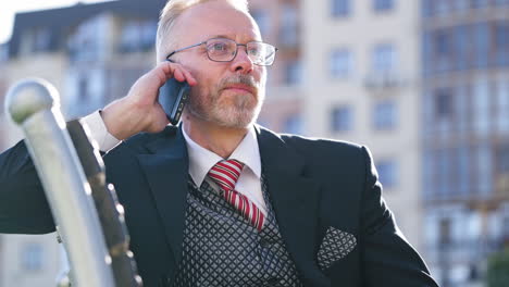 happy elderly businessman talking on smartphone. mature bearded man in suit and glasses sitting on bench and having a conversation about his business in the city.