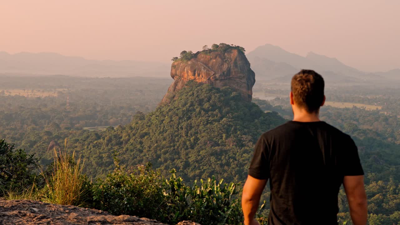 A man gazes thoughtfully at the iconic Sigiriya Rock as the sun rises over the lush landscape of Sri Lanka.
