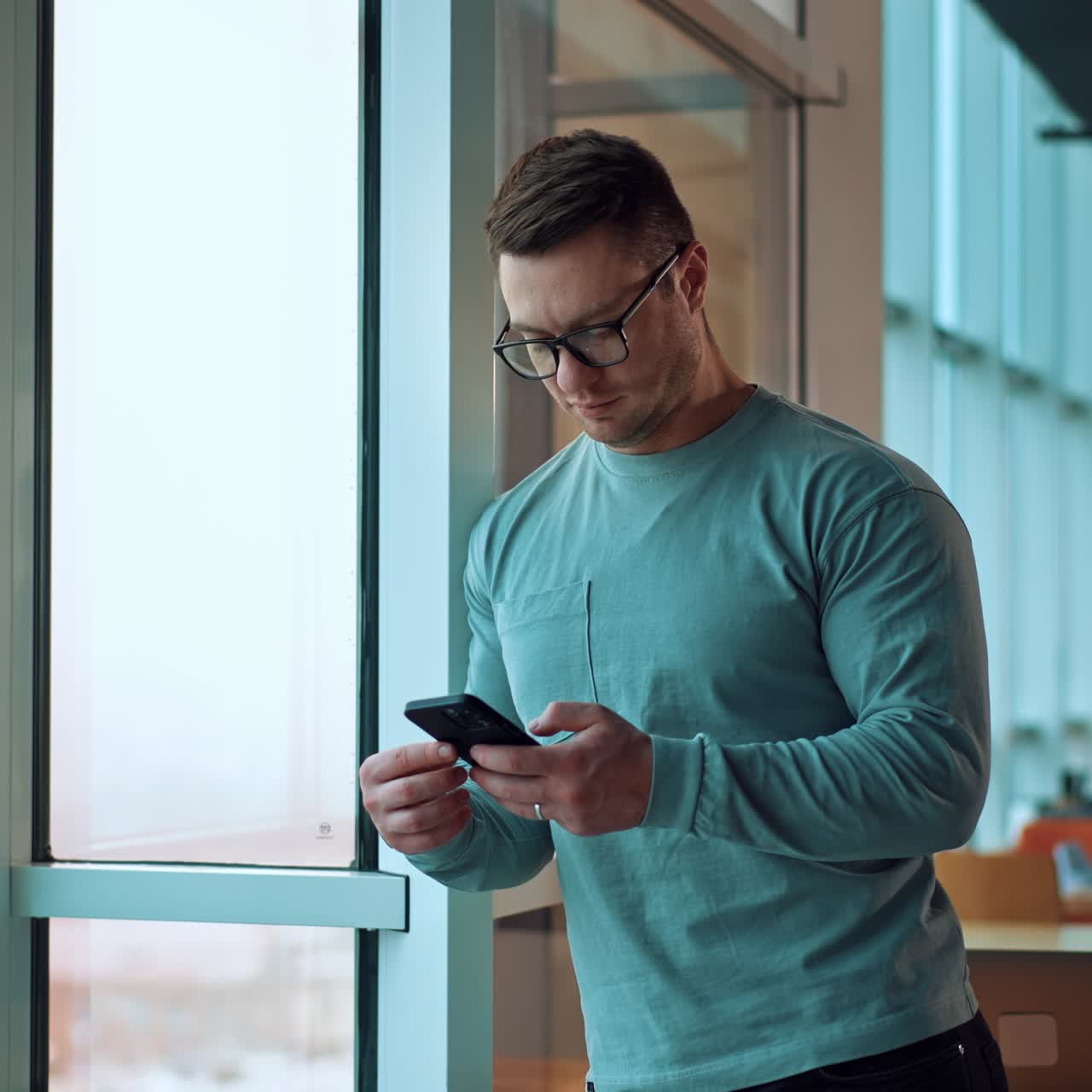 Fit strong dark-haired Caucasian man stands leaning on the window frame. Man in glasses focused on the phone he's holding in his hands