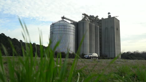 un silo de grano se eleva sobre el campo cercano en primavera mientras los coches pasan - vista deslizante, de ángulo bajo con hierba en primer plano