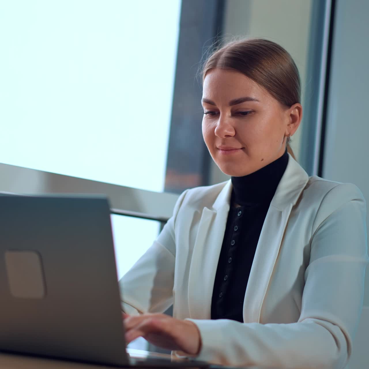 Calm Caucasian lady sitting at the desk working on laptop. Business woman typing on computer low angle view. Window at backdrop