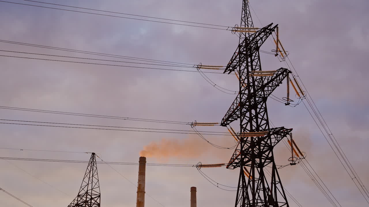 Electricity pylon on industrial chimney background. High-voltage tower and pipe of power plant with smoke on evening sky. Harmful smoke polluting the environment.