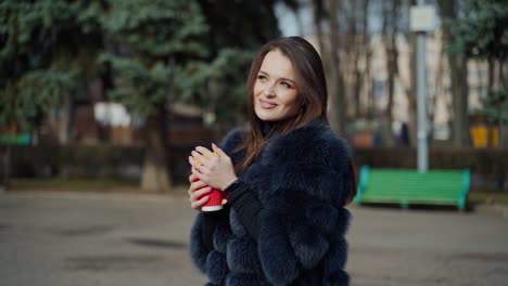 Positive young woman in the park. Attractive girl in stylish fur coat holds plastic cup of coffee and posing on camera outdoors. Slow motion.