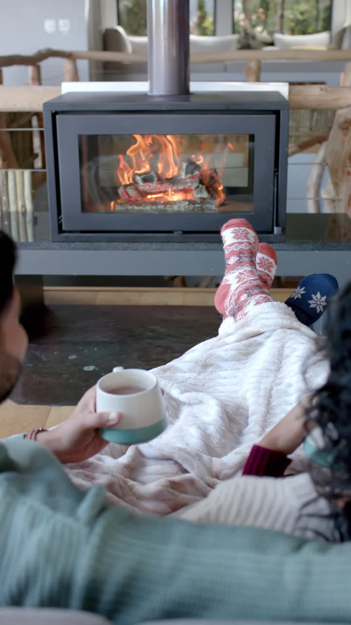 Vertical video of biracial couple sitting on sofa in front of fireplace at home, slow motion