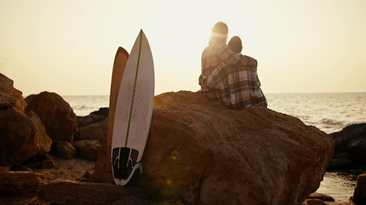vista trasera de un tipo rubio en una camisa a cuadros y su novia en un sombrero negro, girando la camisa, sentado y abrazando en la piedra grande en una orilla rocosa por el mar, cerca de ellos hay sus tablas de surf blancas y amarillas al amanecer en la mañana