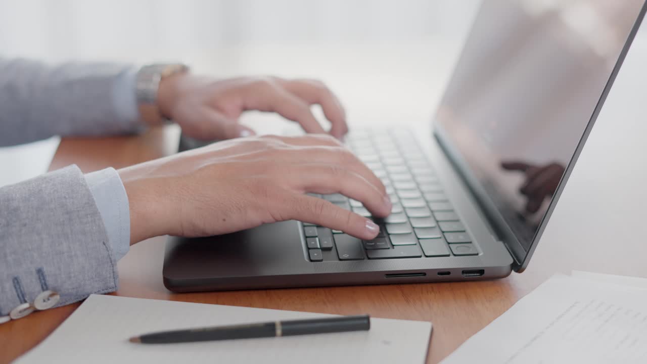 Zoom out shot of hands typing on a laptop keyboard on a desk in a bright office environment