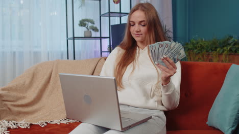 smiling happy girl counting money cash and use laptop pc calculate domestic income earnings at home