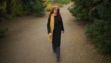 confident lady in a black coat, wearing a yellow beret and matching muffler, looks up and focuses her attention on something intriguing in the background of a serene park